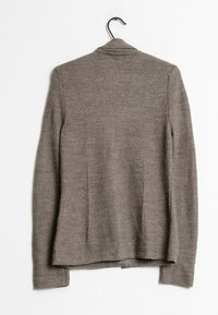 Blazer gris texturizado con mangas largas, ajuste entallado y un patrón sutil, colgado en un gancho negro sobre un fondo blanco.