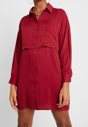 Femme portant une robe chemise rouge ample à manches longues avec des boutons sur le devant et une poche poitrine, accessoirisée avec des colliers en or et des boucles d'oreilles anneaux.