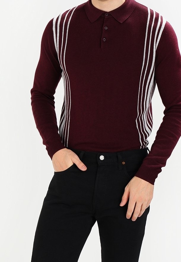 Homme portant un pull polo manches longues bordeaux avec des rayures verticales blanches et un pantalon noir, une main dans la poche, sur fond blanc.