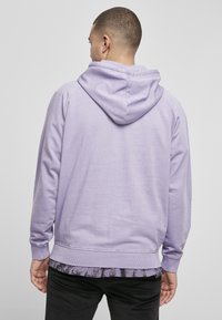 Sudadera de color púrpura claro con capucha ajustable, puños acanalados y dobladillo. Textura de tela suave. Lleva una camisa de patrón negro y gris debajo.