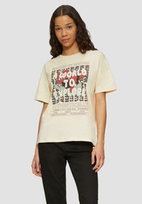 Crème-kleurig oversized t-shirt met een grafisch ontwerp van tekst en abstracte vormen in rood en groen. Korte mouwen en ronde halslijn.