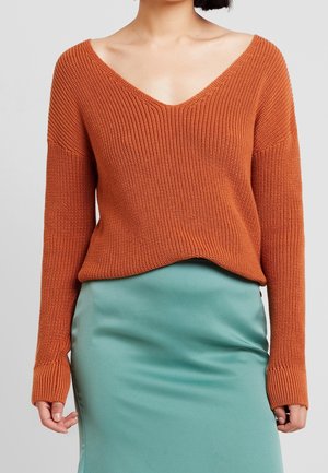 Persona vistiendo un jersey de punto naranja oxidado con un amplio escote en V y una falda de satén azul verdoso, de pie frente a un fondo claro y sencillo.