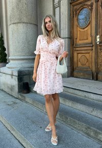 Femme en robe fleurie rose et sandales blanches tenant un sac à main blanc en perles sur des marches en pierre près d'une grande porte en bois.
