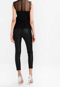 Schwarzes ärmelloses Oberteil mit Spitzen- und Pünktchen-Mesh-Details, kombiniert mit figurbetonten schwarzen Lederimitat-Hosen und silbernen High-Heels.