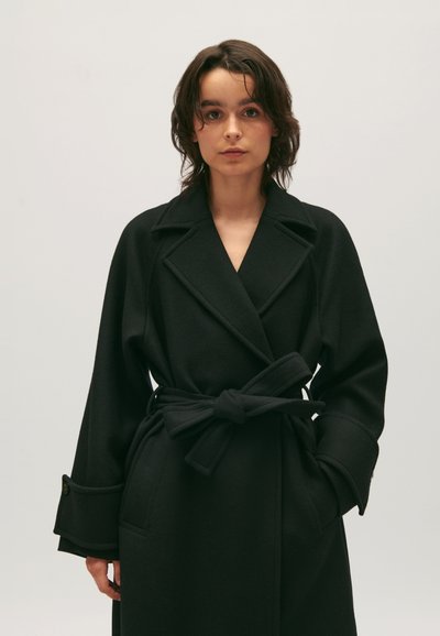 Zwarte wollen trenchcoat met een sjawlkragen, een ceintuur in de taille, diepe zakken en brede mouwen. Gelaagd stof en strakke lijnen.