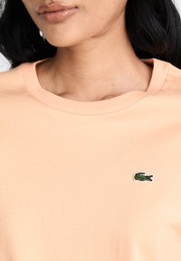 Pfirsichfarbener Baumwoll-T-Shirt mit Rundhalsausschnitt und kurzen Ärmeln, versehen mit einem kleinen grünen gestickten Krokodil-Logo auf der linken Brust.