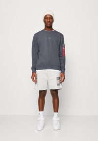 Szary sweatshirt z kontrastowym przeszyciem, czerwoną kieszenią na suwak na rękawie, w połączeniu z jasnoszarymi szortami i białymi trampkami. Na głowie kremowa czapka.