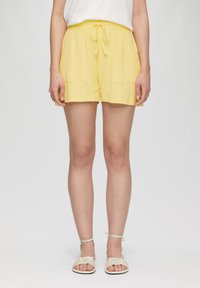 Shorts jaunes avec taille élastique et cordon de serrage, deux poches avant et un tissu texturé, associés à des sandales beiges à lanières.