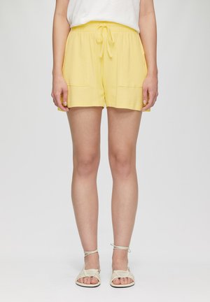 Gele, elastische broek met een koord, twee voorzakken en een gestructureerde stof, gecombineerd met beige sandaaltjes met banden.