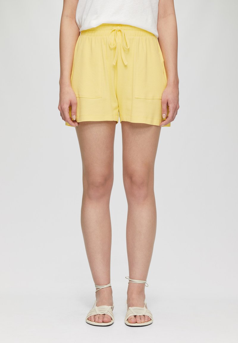 Shorts jaunes avec taille élastique et cordon de serrage, deux poches avant et un tissu texturé, associés à des sandales beiges à lanières.