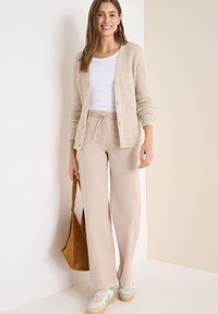 Beige ribbed cardigan päällä valkoisen paidan, vaaleanbeige leveälahkeiset housut ja kermaiset lenkkarit sinisillä yksityiskohdilla. Ruskea olkalaukku kädessä.