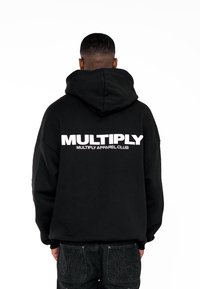 Felpa nera con cappuccio con logo "MULTIPLY" in bianco sulla schiena, caratterizzata da una vestibilità rilassata e polsini a coste. Indossata con jeans scuri.