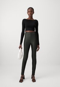 Crop top noir à manches longues ; leggings taille haute verts foncés brillants ; sandales à talons carrés noires ; sac pochette blanc. Textures lisses, design ajusté.