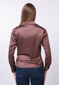 Καφέ σατέν πουκάμισο με γιακά, μακριά μανίκια και λεπτομέρειες με κουμπιά στις μανσέτες. Φορεμένο με σκούρο μπλε τζιν, που αποκαλύπτει την πίσω όψη.