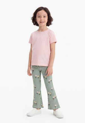 Enfant debout portant une chemise rose pâle à manches courtes, un pantalon vert à motif floral avec des ourlets froncés, et des chaussures blanches à enfiler.