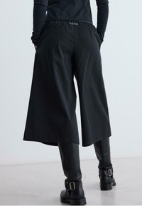 Person trägt weite schwarze Hosen über schmalen schwarzen Stiefeln mit Schnallen und steht mit dem Rücken zu einem hellgrauen Hintergrund.