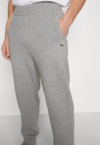 Šedé tepláky vyrobené z měkké tkaniny, s elastickým pasem a bočními kapsami, s malým logem Lacoste umístěným poblíž kapsy.