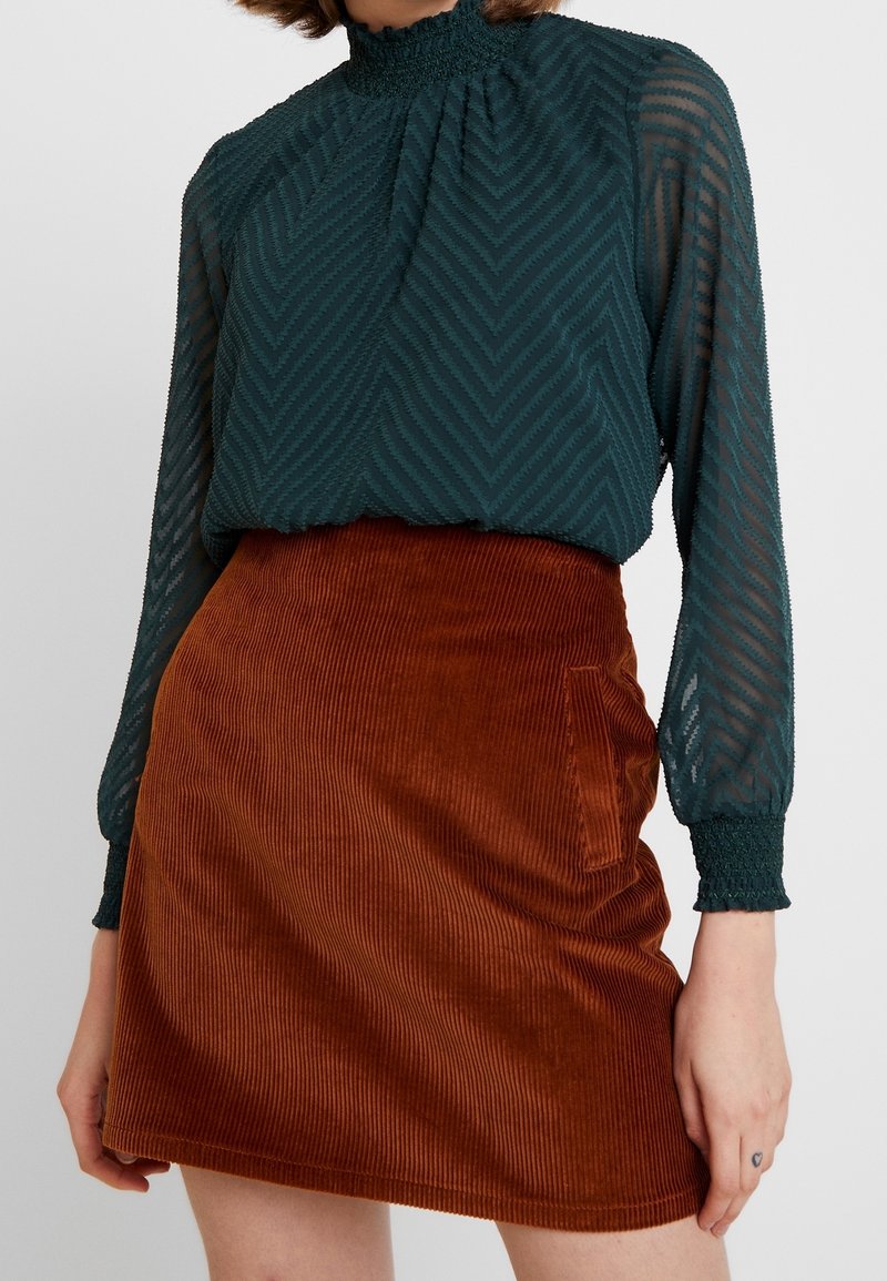 Blouse teal à motifs avec un col haut froncé et des manches longues ; associée à une mini-jupe en velours côtelé couleur rouille dotée d'une poche latérale.