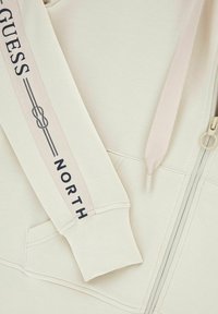 Sweat à capuche beige zippé avec poche avant, cordons de serrage, et texte noir "GUESS NORTH" accompagné d'un motif de corde sur la manche.