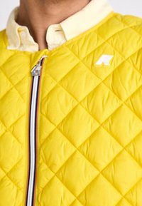 Primo piano di una persona che indossa una giacca trapuntata gialla con una cerniera a strisce multicolori e una camicia colletto giallo pallido sotto.