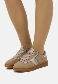 Beige Wildledersneaker mit Gummisohle und flachen Schnürsenkeln, die ein seitliches Paneeldesign und Logoakzente aufweisen.