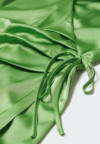Tissu en satin vert avec détail froncé et longues lanières fines drapées sur une surface lisse, mettant en valeur la texture et la brillance.
