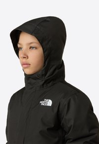 Giacca nera con cappuccio dalla texture liscia, con un logo bianco sul petto. Il design è funzionale con cappuccio aderente.