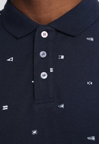 Polo bleu marine avec motifs de drapeaux nautiques blancs, patte de boutonnage et texture de tissu doux. Présente un col classique.