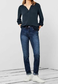 Femme portant un chemisier bleu marine à manches longues, un jean en denim retroussé et des baskets blanches montantes, debout les mains dans les poches.