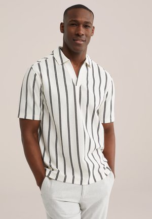Homme debout les mains dans les poches, portant une chemise blanche à manches courtes avec des rayures verticales noires et un pantalon gris clair.