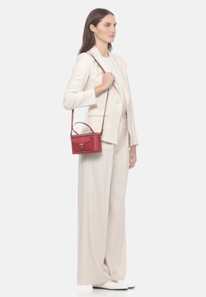 Donna in completo crema che tiene una piccola borsa rossa con manico superiore e tracolla, in piedi davanti a uno sfondo semplice.