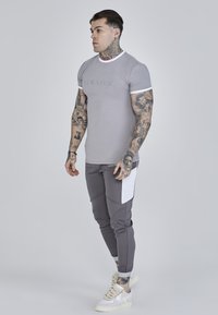 Grå tætsiddende t-shirt med hvid krave og logo, sammen med grå tapered bukser med en hvid indsatslomme og hvide sneakers.