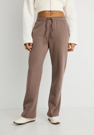 Personne portant un pantalon de survêtement marron à cordon, une veste courte blanche et des baskets blanches, debout devant un fond clair uni.