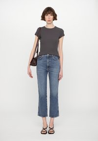 Mörkgrå tajt T-shirt, högmidjade blå jeans med fransad fåll och svarta sandaler med remmar. Modellen håller en liten brun handväska.