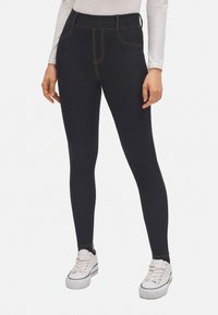 Leggings azul oscuro con cintura alta, con costuras amarillas, textura suave y corte ajustado. Llevados con zapatillas blancas.