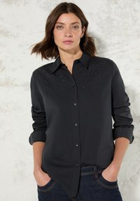Camicia nera con colletto, caratterizzata da borchie decorative sulle spalle. Le maniche lunghe sono arrotolate e il tessuto sembra liscio.