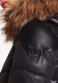 Sort pufferjakke med en faux fur-krave, der har et tekstureret syntetisk stof, quiltet design og en mærket sort detalje på skulderen.