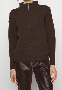 Femme portant un pull en tricot marron foncé avec une fermeture éclair argentée à l'avant et un pantalon en cuir marron foncé avec des panneaux latéraux et des coutures blanches.