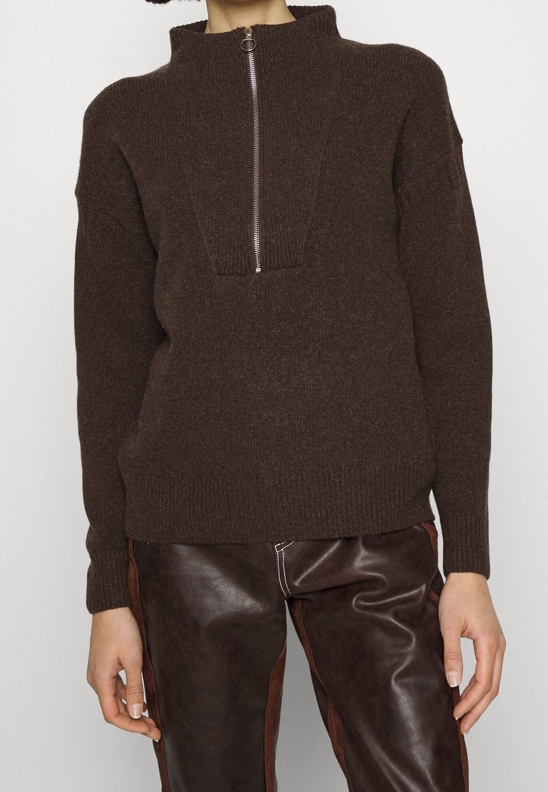 Femme portant un pull en tricot marron foncé avec une fermeture éclair argentée à l'avant et un pantalon en cuir marron foncé avec des panneaux latéraux et des coutures blanches.