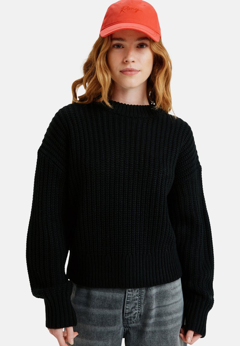Black knitted sweater with a ribbed texture and crew neckline, paired with an orange cap featuring embroidered detailing, worn casually.