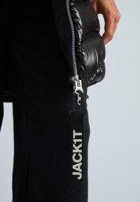 Melna puffer jaka ar spīguļojošu, kviltu struktūru un sudraba rāvējslēdzēju; tajā ir melns siksniņas sīkrīks ar baltu tekstu "JACK1T".