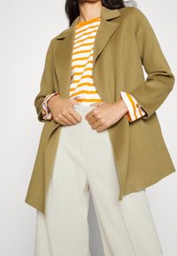 Cappotto in misto lana verde oliva con collo a punta, indossato sopra un top bianco e arancione a righe, abbinato a pantaloni beige chiaro a vita alta.