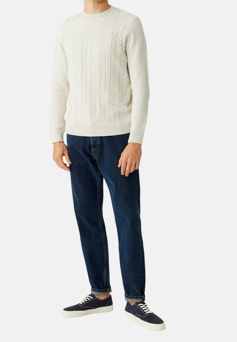 Pull crème en maille torsadée avec col rond, assorti à un jean bleu foncé et des baskets bleu marine à semelles blanches. Look simple, texturé et décontracté.