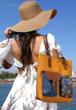 Tan leather tote bag with large orange body, brown accents, and blue floral pocket. Model wears a white, off-shoulder dress and wide-brim hat.