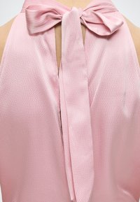 Ροζ τοπ χωρίς μανίκια με υφή και μεγάλο φιόγκο δεμένο στο ντεκολτέ, που δείχνει μια μικρή σχισμή κάτω από τον φιόγκο.