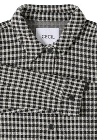 Czarna i biała koszula w szachownicę z klasycznym kołnierzykiem, zapięciem na guziki i metką. Tkanina ma teksturę z wzorem w kratkę na całej powierzchni.