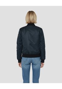 Blouson aviateur bleu marine avec col et poignets côtelés. Tissu lisse et brillant avec une poche latérale. Porté avec un jean bleu clair.