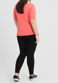 Kvinna står med en korallfärgad t-shirt med korta ärmar, svarta leggings och svartvita sneakers, mot en ljus enfärgad bakgrund.