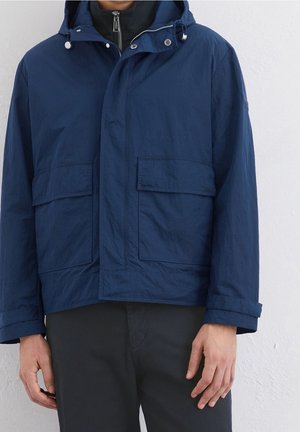 Mężczyzna w granatowej kurtce z kapturem z przednimi kieszeniami z patkami i czarnych spodniach na tle jasnej ściany.