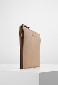 Úzká béžová kožená kabelka s zlatým zipem a malým zlatým logem, stojící vzpřímeně na bílé ploše na jednoduchém pozadí.